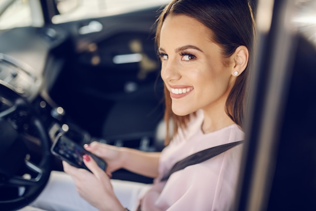 Gorgeous cheerful young Caucasian brunette sitting in car with safety belt on and typing message.