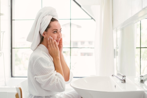 Великолепная уверенная в себе взрослая женщина с полотенцем на голове, сидящая и проверяющая свое лицо перед зеркалом в домашней ванной комнате Довольно привлекательная женщина, образ жизни красоты и косметическая концепция