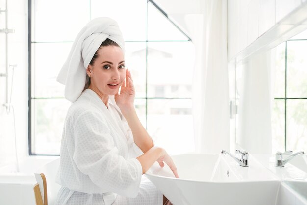 Gorgeous confident adult woman wearing a towel on her head sitting and checking her jaw in front of the mirror in the home bathroom A pretty attractive woman beauty lifestyle and cosmetic concept