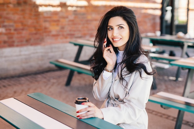 Gorgeous darkhaired woman with pure skin dark bright eyes and full lips holding takeaway coffee and smartphone chatting with her boyfriend having pleased look Young female enterpreneur resting