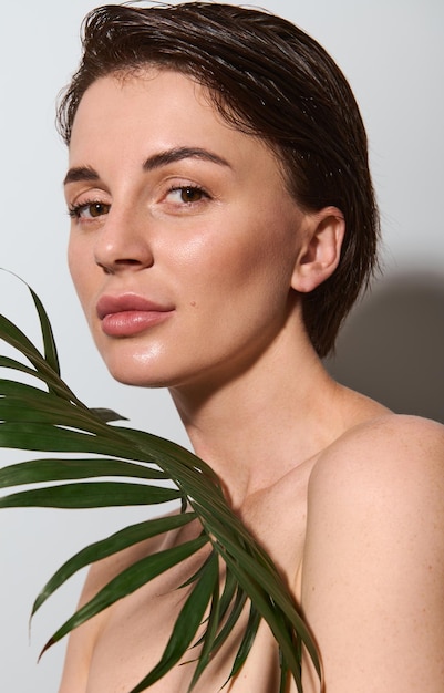 Photo gorgeous delightful beautiful young brunette of natural beauty poses nude with palm leaf on a white background. skin and body care, wellness and well-being concept