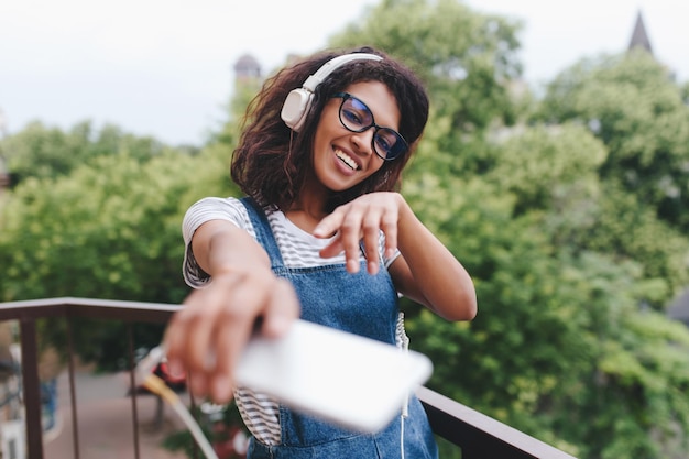 Gorgeous enthusiastic girl with short curly hair dancing on balcony in morning and laughing. Outdoor portrait of pleased young woman in headphones with smartphone having fun and smiling.