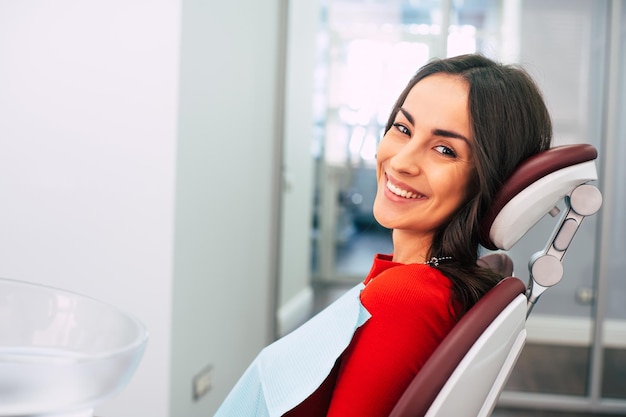 La splendida ragazza che indossa un maglione rosso nella stanza di stomatologia piena di luce diurna e colori bianchi sta sorridendo con il suo nuovo sorriso bianco accattivante