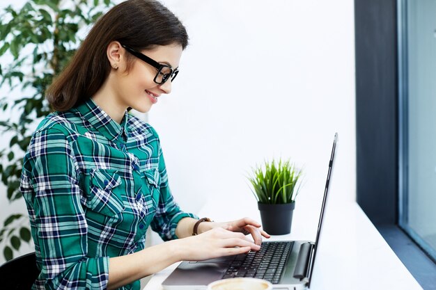 Gorgeous girl with black hair wearing shirt and eyeglasses sitting in cafe with laptop and cup of coffee, freelance concept, working process.