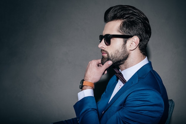 Gorgeous and handsome. Close-up side view of young handsome man in suit and bow tie holding hand on chin and looking away while sitting against grey background