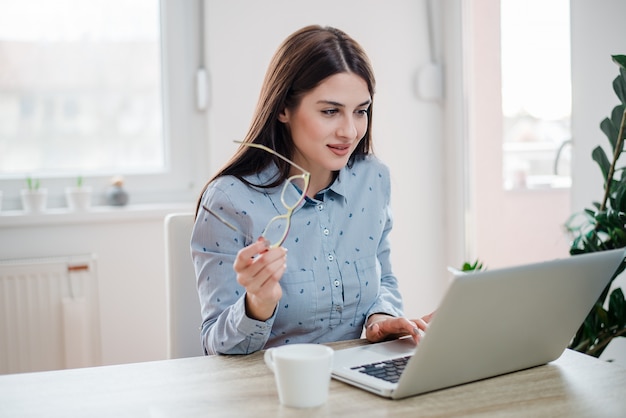 Gorgeous long haired woman holding eyeglasses while using laptop. Freelance work concept.