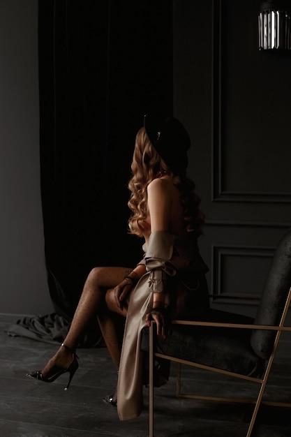 A gorgeous model girl with long sexy legs wearing stockings, high heels shoes, trench coat, and hat poses into the dark vintage interior