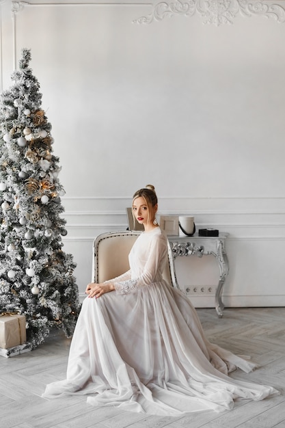 A gorgeous model woman in a fashionable dress sits near the Christmas tree in the vintage interior