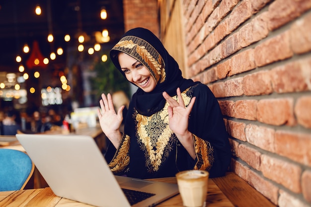 Gorgeous muslim woman in traditional wear having video call over the laptop while sitting in cafeteria and drinking coffee.