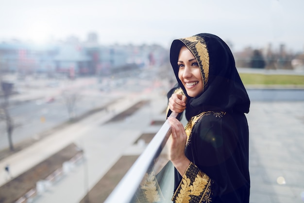 Gorgeous muslim woman in traditional wear looking at beautiful sight while standing on rooftop.