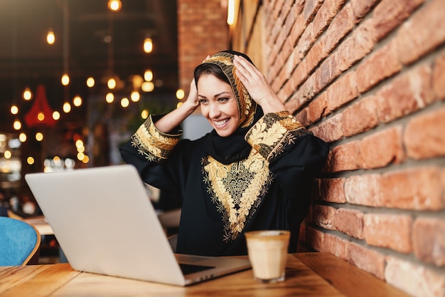 Gorgeous muslim woman with toothy smile dressed in traditional wear using laptop for on-line shopping while sitting in cafeteria. On desk coffee.