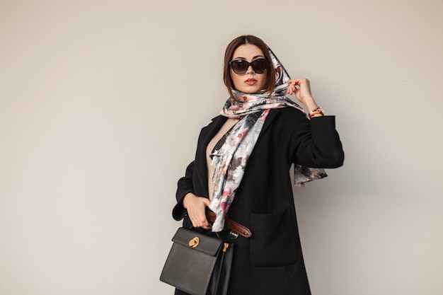 Gorgeous pretty young professional woman in stylish sunglasses in trendy black coat with leather bag with a vintage scarf on head posing near a wall outdoors. Attractive girl fashion model. Sexy ladie