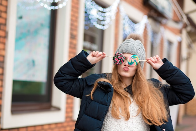 灰色のニット帽をかぶって、店の窓の近くでカラフルなクリスマスのキャンディーを保持しているゴージャスな赤い髪の若い女性