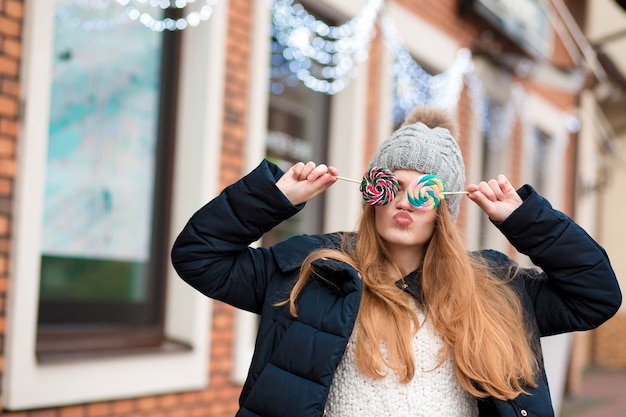 Splendida giovane donna dai capelli rossi che indossa un cappello lavorato a maglia grigio e tiene in mano caramelle colorate di natale vicino alla vetrina del negozio