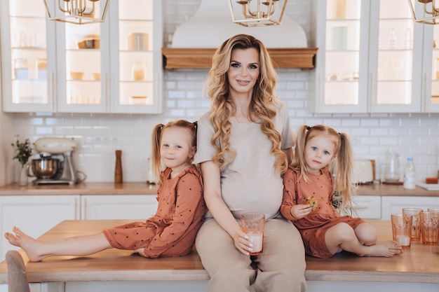 Gorgeous Scandinavian pregnant woman in beige casual clothes sits on desk with two little daughters