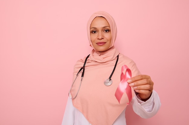 Splendida dottoressa araba musulmana serena in hijab rosa che tiene nastro di raso rosa, guardando la fotocamera, isolata su sfondo colorato, copia spazio. giornata mondiale della lotta contro il cancro al seno, concetto del 1° ottobre