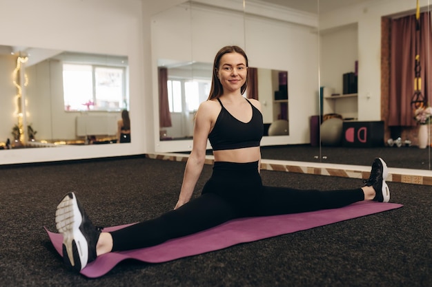 Великолепная стройная девушка, одетая в черный спортивный топ и колготки, делает растяжку на коврике для фитнеса в тренажерном зале Yoga Fitness Sport