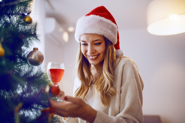 Gorgeous smiling caucasian blonde woman with santa hat on touching ornament on christmas tree and drinking wine while standing in living room.