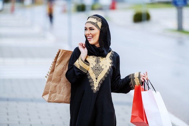 伝統的な服装でゴージャスな笑顔の肯定的なアラブの女性が通りを歩いて、彼女の買い物に満足していると感じて買い物袋を手に持っています。