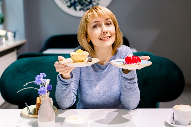 Giovane donna sorridente splendida che mangia dolce e che beve caffè ad un self-service. donna con due piatti di torte seduti al tavolo