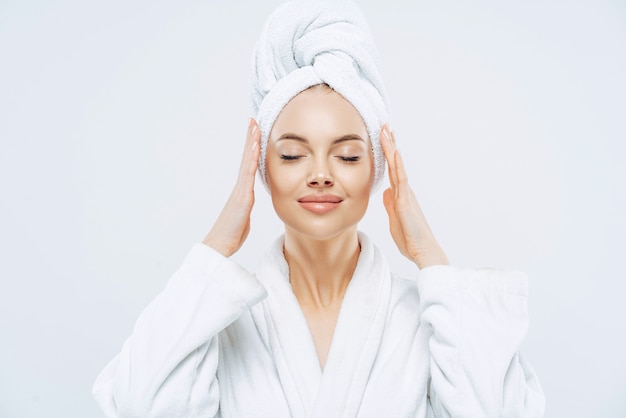 Gorgeous spa woman stands with closed eyes, keeps hands on towel wrapped on head, dressed in white bath robe, has healthy skin, natural makeup, well groomed complexion, poses indoor. beauty treatment