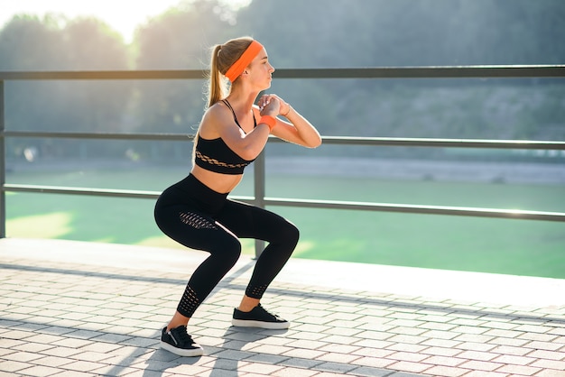 Ragazza sportiva splendida in abiti sportivi neri e fascia arancio che squating durante l'allenamento di forma fisica allo stadio all'aperto.