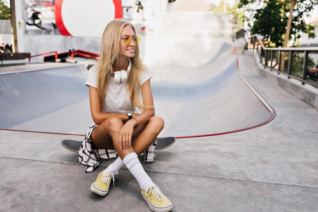 Gorgeous tanned skater girl posing outdoor Photo of smiling female model in sport shoes sitting with legs crossed on skateboard