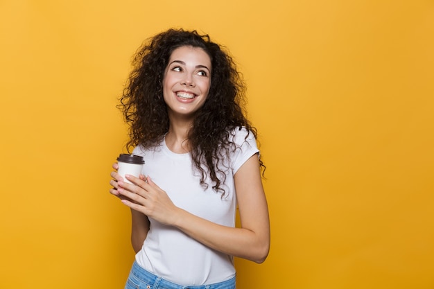 gorgeous woman 20s with curly hair smiling and holding takeaway coffee in paper cup isolated on yellow