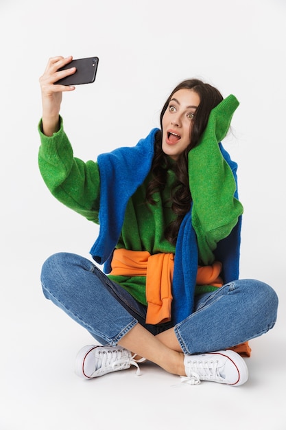 gorgeous woman in colorful clothes sitting on the floor and taking selfie on smartphone, isolated on white