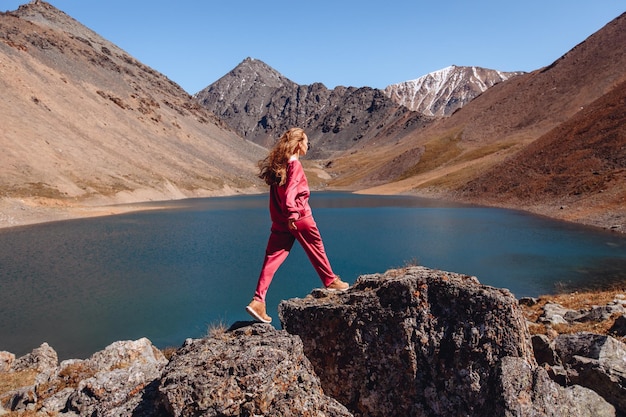 A gorgeous woman in a red suit walks on cobblestones next to a tiny reservoir in the Rocky Mountains