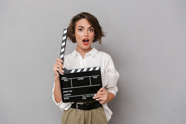 gorgeous woman smiling and holding black clapperboard, isolated over gray wall