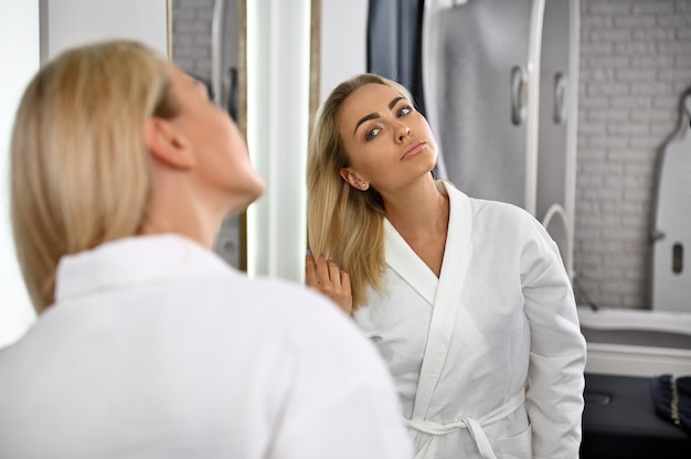 A gorgeous woman in a white robe touches her blonde hair, admiring her reflection in the mirror. Beauty portrait of attractive woman with perfect smooth skin