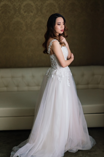 Gorgeous young asian woman in elegant dress looking at camera and window, morning of the bride, wedding