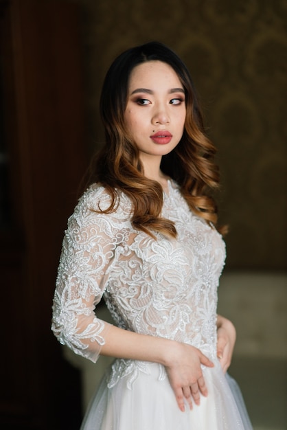 Gorgeous young asian woman in elegant dress looking at camera and window, morning of the bride, wedding