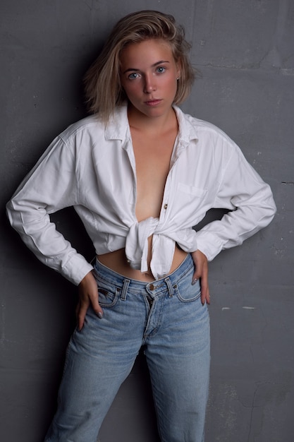 A gorgeous young blonde woman in an unbuttoned shirt and jeans poses on a gray background