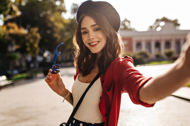 Gorgeous young brunette with medium wavy hair red lips beret white blouse and red shirt smiling and making selfie against sunlit city background