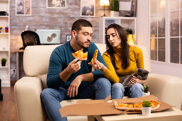 ソファに座ってリビングルームでテレビを見ながらピザを食べるゴージャスな若いカップル