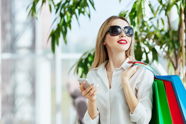 Gorgeous young girl with light brown hair and red lips wearing white blouse and standing with colorful shopping bags, holding mobile phone, shopping concept, copy space.
