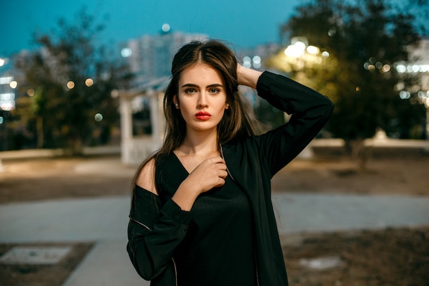 Шикарная молодая модельная женщина смотря камеру представляя в городе нося черное платье вечера.