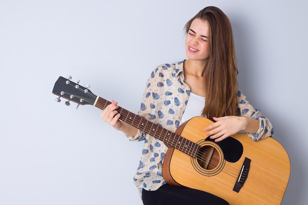 Gorgeous young woman in beige blouse and black trousers playing the  guitar on gray background in studio