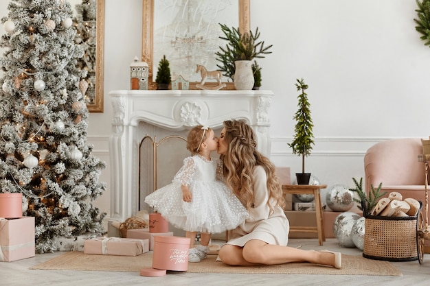 Gorgeous young woman in stylish dress posing with her little daughter in fluffy dress in Christmas decorated interior.