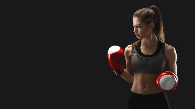 Gorgeous young woman with boxing gloves, standing in the defending position, ready to fight, copy space. Studio shot on black background, low key. Kickboxing and fight sport concept