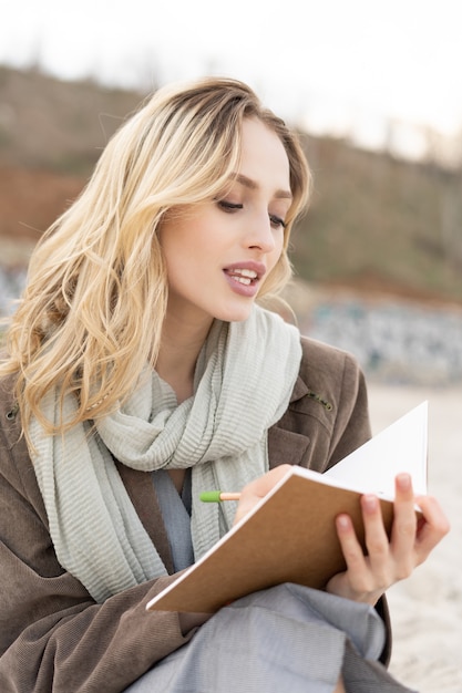 Gorgeous young woman writing in the notebook sitting on the beach with a stylish outfit