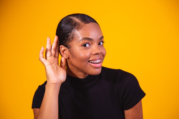 Gossip concept. Curious young afro woman holding hand near ear, trying to hear interesting information, cheerful korean female eavesdropping on yellow studio background, panorama with copy space.