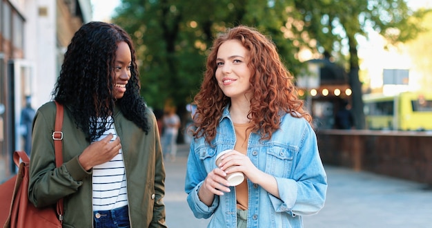 Gossip girls. Waist up portrait view of the two multiracial girlfriends chatting and drinking coffee while walking at the summer street. Vacations concept