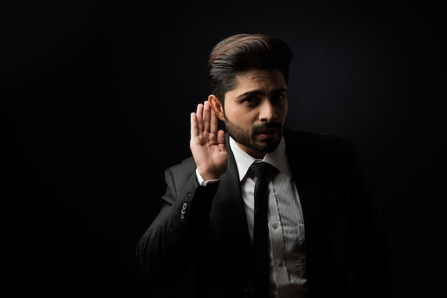 Сплетни или слухи - индийский бизнесмен слушает с позой подслушивания на черном фоне