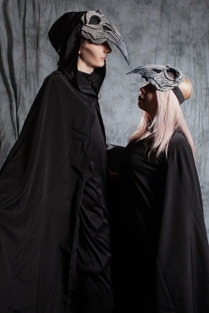 A gothic couple dressed as ravens a black cape with a hood and a bird mask