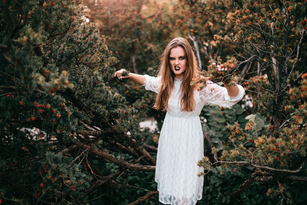 Gothic girl in white vintage dress posing against bushes in summer park.