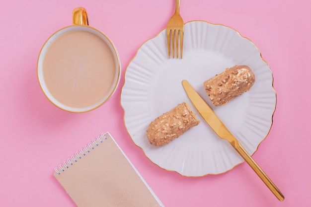 Gouden gekleurde eclairs op plaat met mes en vork met kopje koffie op roze achtergrond