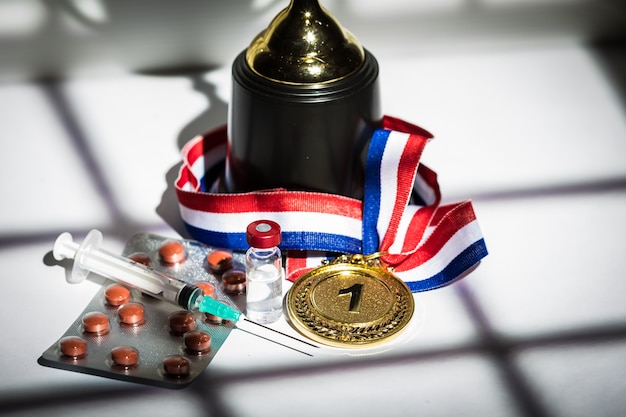 Gouden medaille en kampioensbeker, spuit met dopingmiddel, piltablet en flacon met verboden stof met lichten en schaduwen van een gordijn dat door het raam binnenkomt. Sport- en dopingconcept